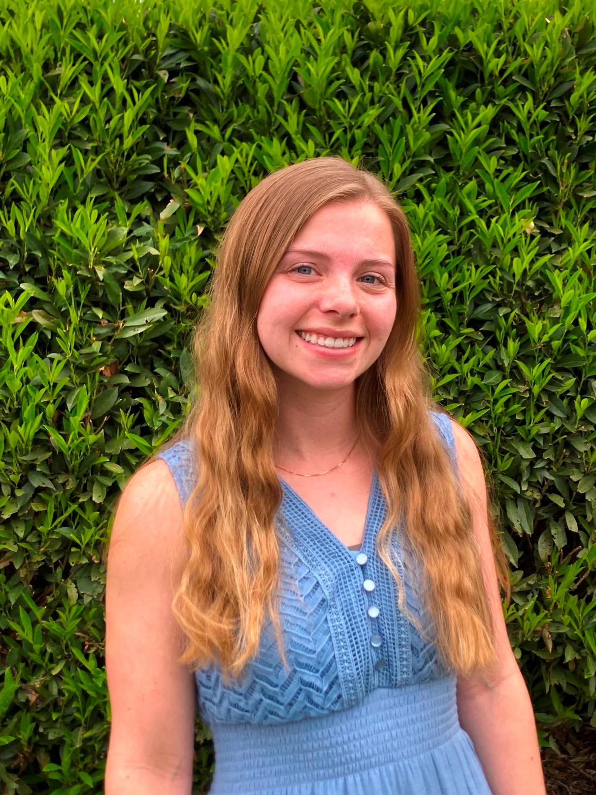 Lexi Bryant, a white woman with long lightly wavy dirty blonde hair, wearing a blue tank top dress. She is standing in front of a hedge and turned slightly to the left.