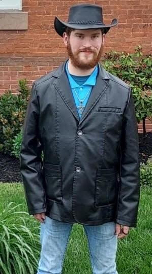 Joseph Scott, a white man with ginger hair and a full beard, wearing a black leather jacket, blue button-down, and black cowboy hat. He is standing in front of a brick building with a manicured lawn and shrubs surrounding it. 