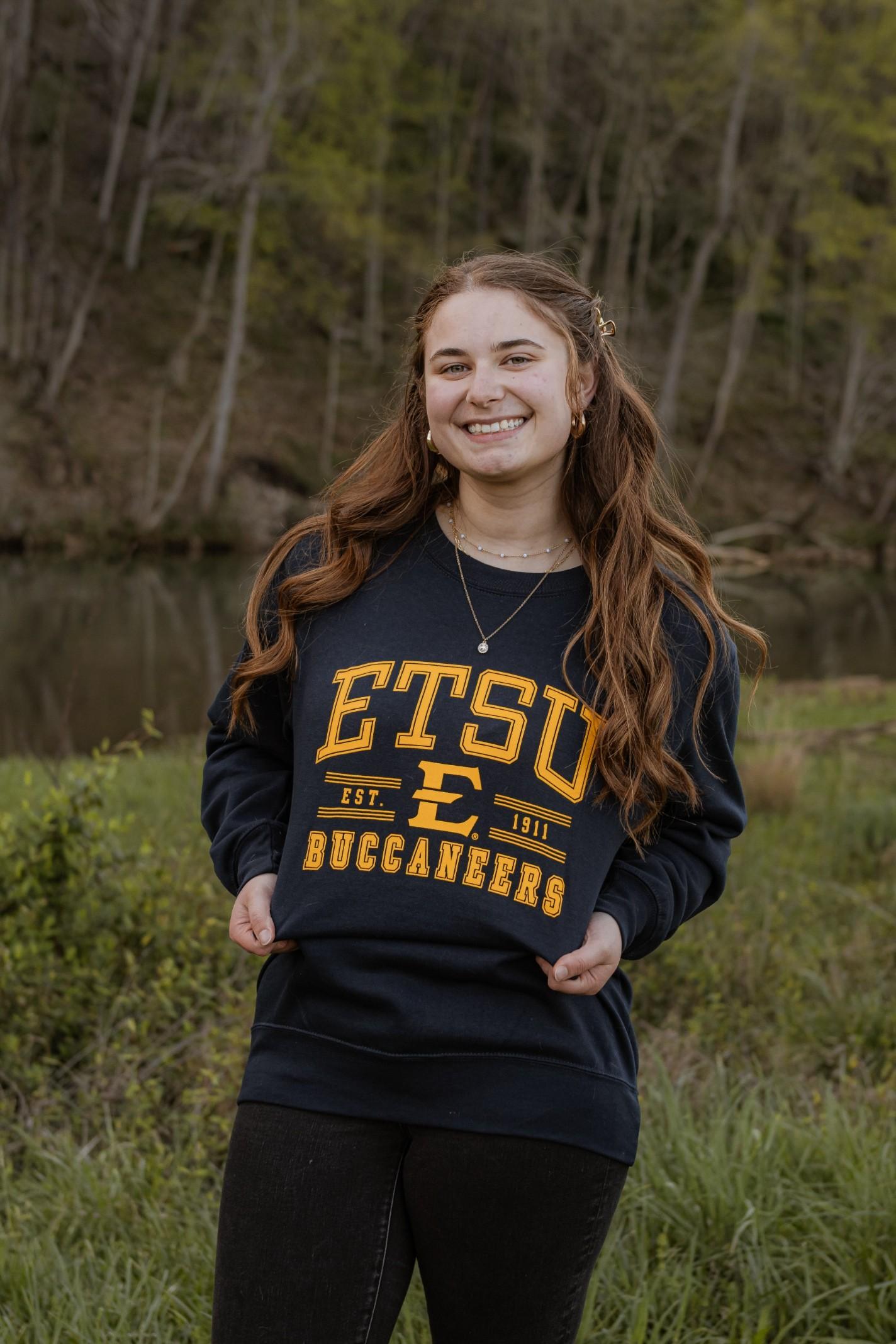 Katherine Mazzei, a white woman with long auburn hair, wearing an ETSU sweatshirt and jeans. She is standing in front of a small river with woods behind it. She is showing off her sweatshirt by pulling it so the text is clearly visible. 