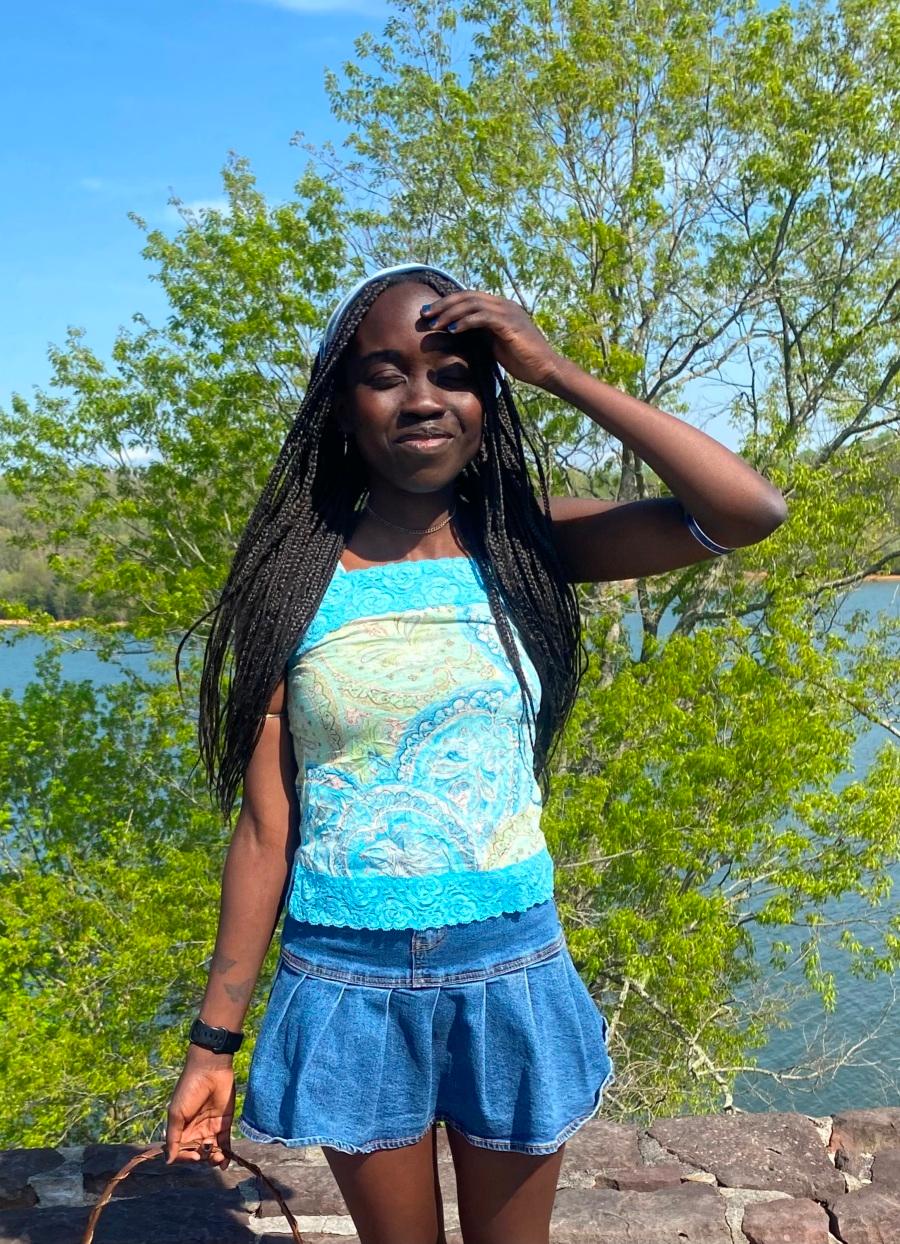 Suzi Peter, a Black woman with long dark braided hair, wearing a blue patterned tank top and blue jean skirt. She is standing in front of a low stone wall with water and a small tree behind her. She is covering her eyes from the sun. 