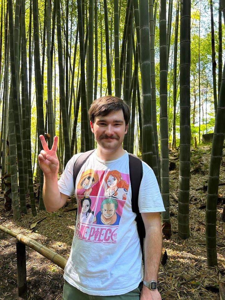Matthew Barker, a white man with brown hair and a mustache, wearing a white One Piece shirt and backpack in front of a thick bamboo thicket holding up a peace sign.