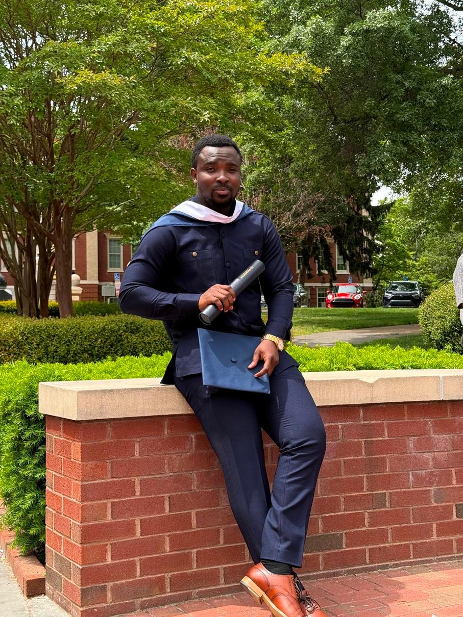 Jerry Kofie, a Black man with short dark brown hair and goatee, wearing graduation regalia leaning on a low brick wall with abundant foliage behind him. He is holding a graduation cap and certificate tube.