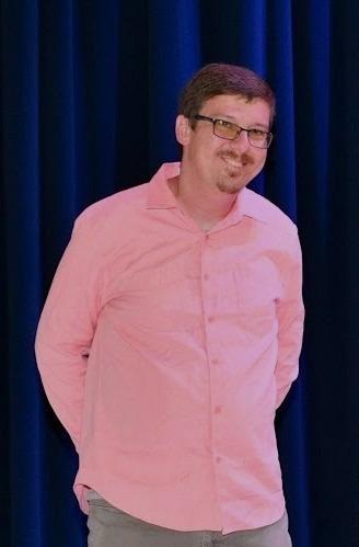 Jonathan Stokely, a white man with short brown hair and a goatee, wearing a pink button down shirt and glasses. He is standing in front of dark blue curtains.