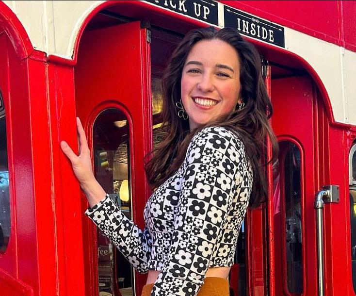 Adeline Rosebush, a white woman with shoulder length dark brown hair, wearing a black and white patterned shirt. She is posing on a red and white double-decker bus.
