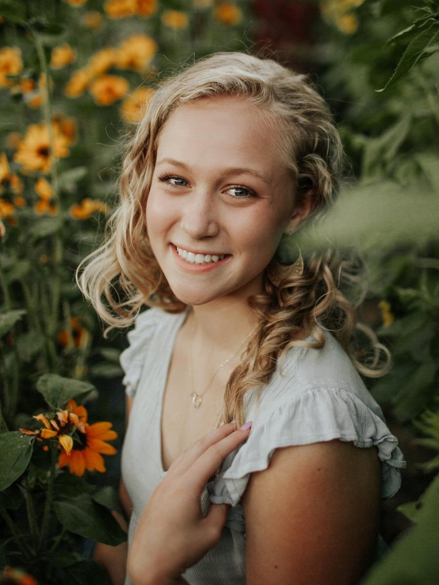 Zayda Carver, a white woman with shoulder length lightly curled blonde hair, wearing a short sleeve gray top and a small gold necklace. She stands in a field of sunflowers looking slightly up at the camera.