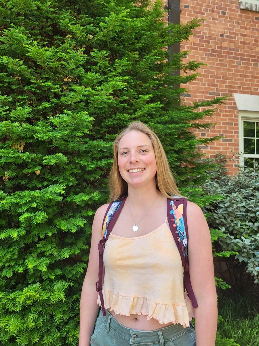 Rachel Brennan, a white woman with long straight dirty blonde hair, wearing a yellow tank top, jeans, and multicolor backpack. She is standing in front of a fur tree and other foliage.