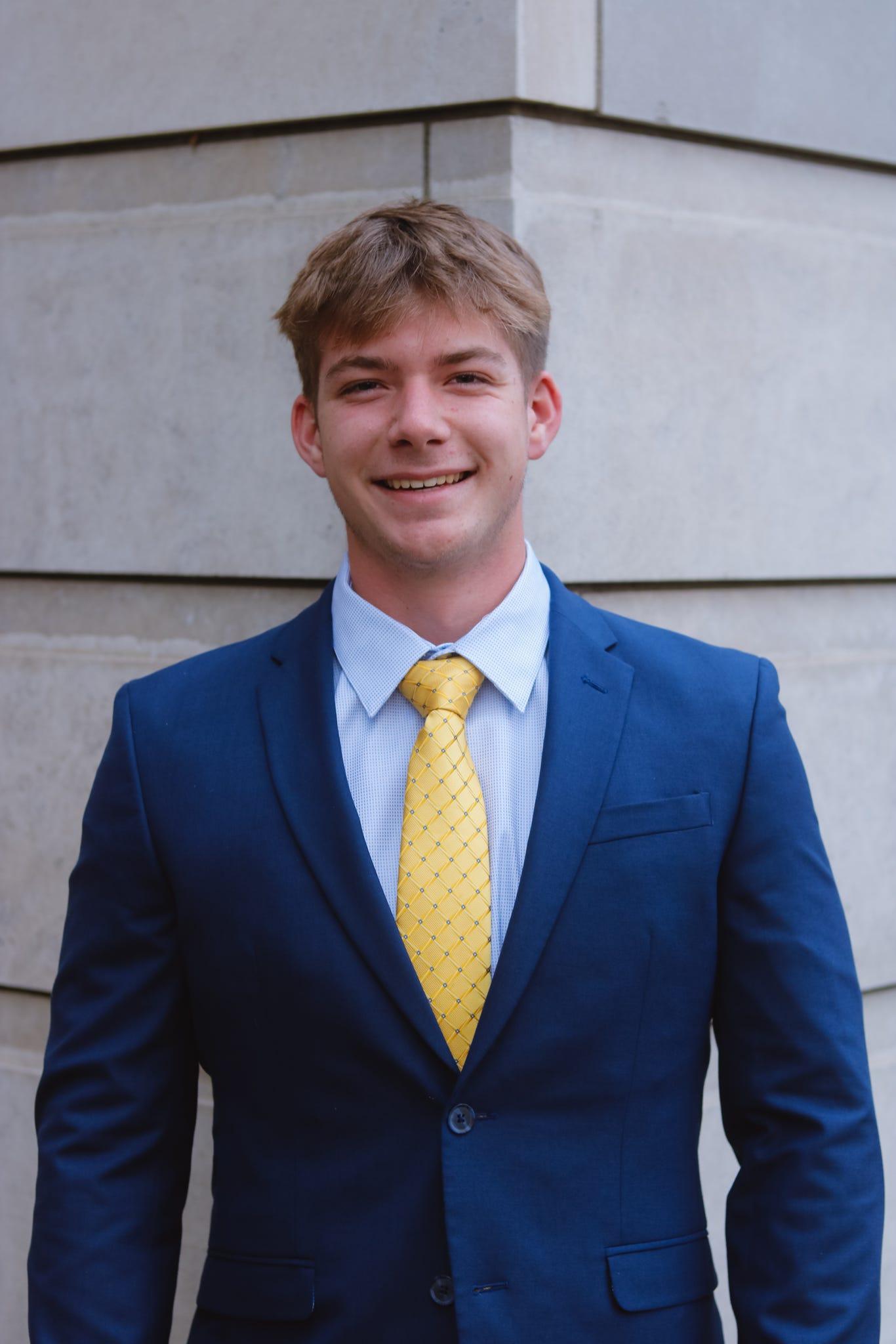 Jared Huff, a white man with short light brown hair and clean shaven face, wearing a two piece blue suit with light blue button down and gold tie. He is standing at a corner of a gray building, facing the camera directly. 