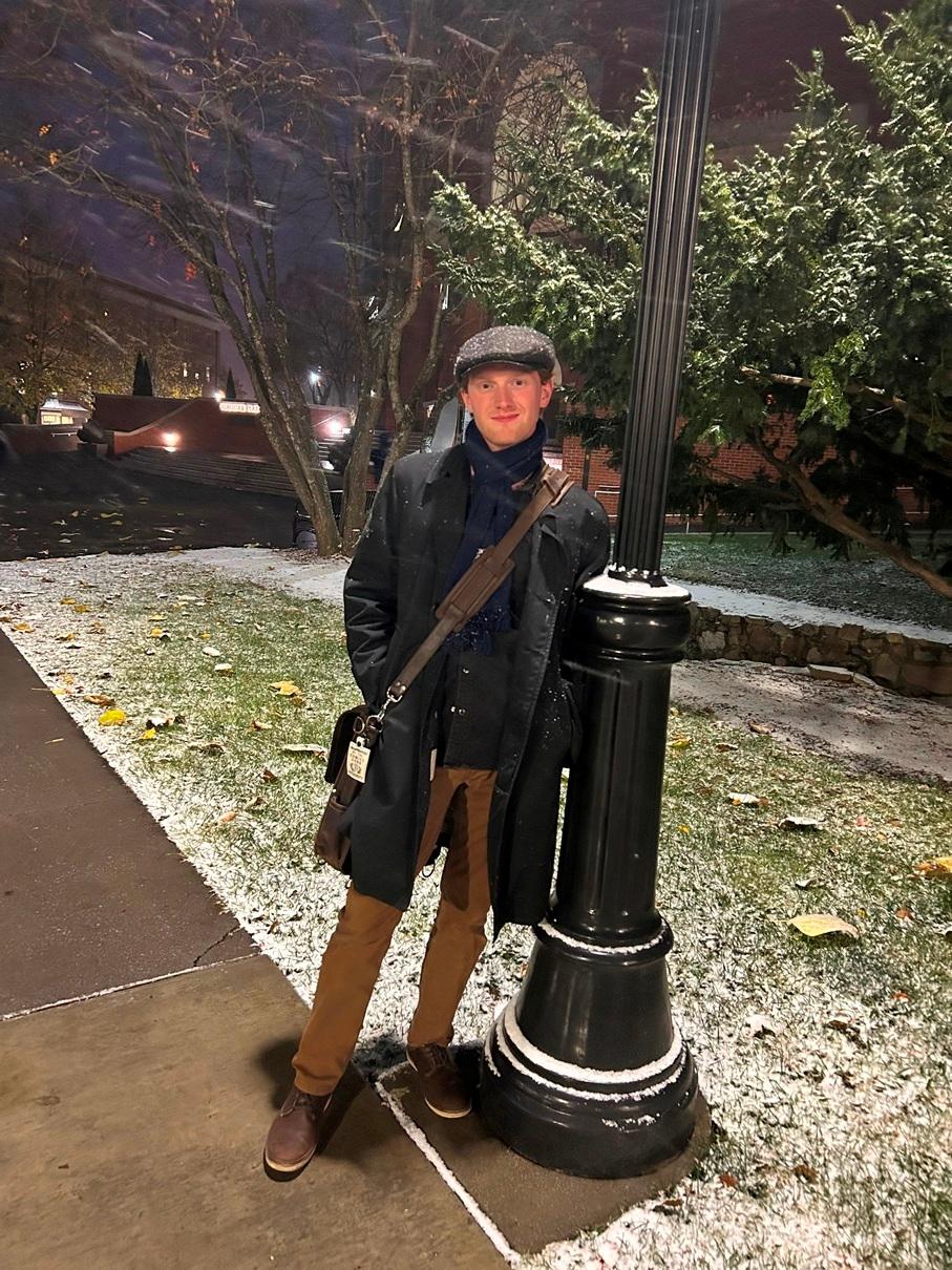 Ben Bilderback, a white man with ginger hair and a clean shaven face, wearing a black overcoat, khakis, a messenger bag, and a newsboy hat. He is leaning on a lightpole in a small dusting of snow.