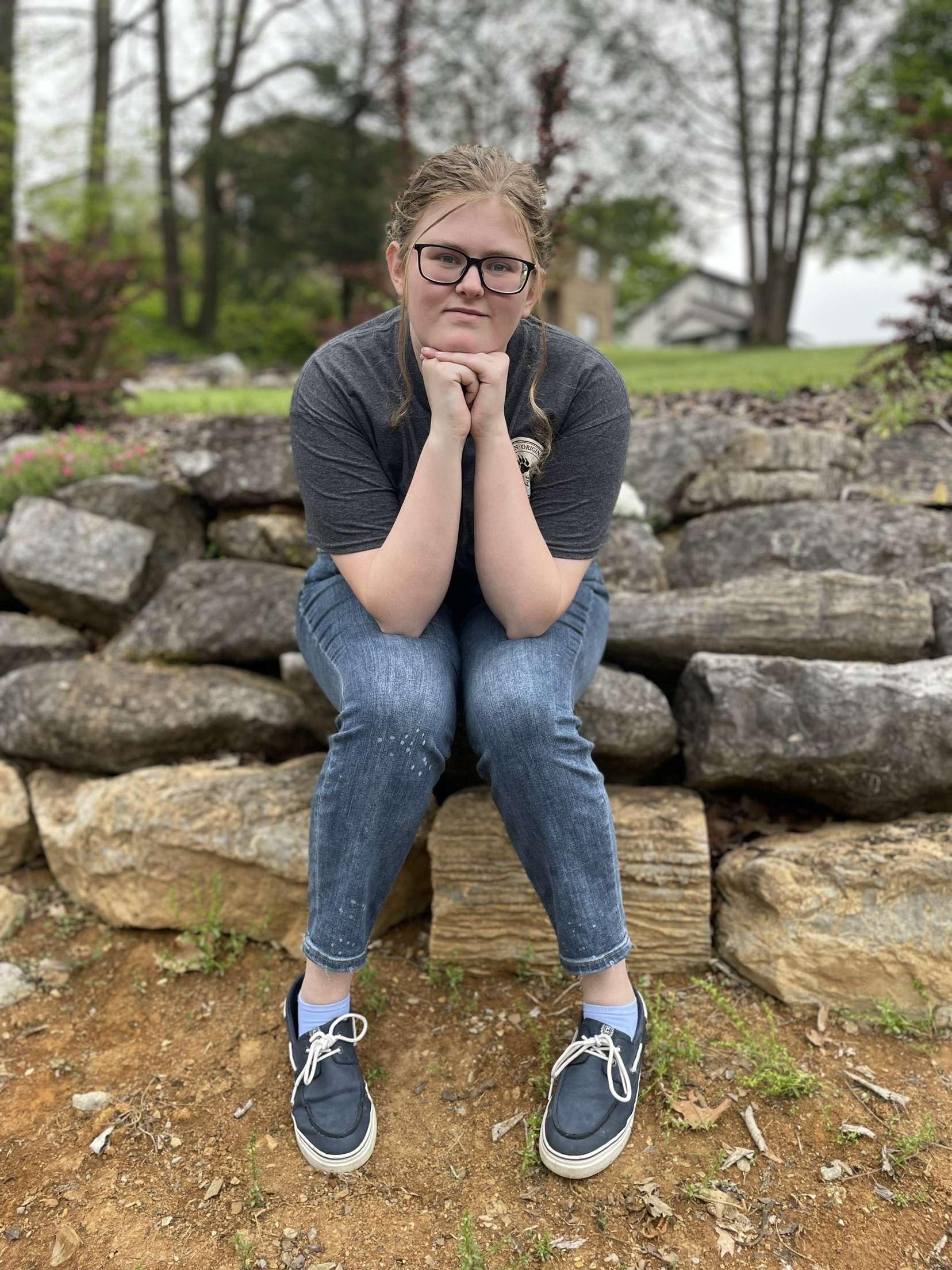 Casey Grindstaff, a white woman with dirty blonde hair bulled into a ponytail, wearing a dark gray t-shirt, jean capris, and blue boat shoes. She is sitting on landscaped rocks with her hands clasped together under her chin and elbows resting on her knees. Behind her are large trees and a house on a hill. 