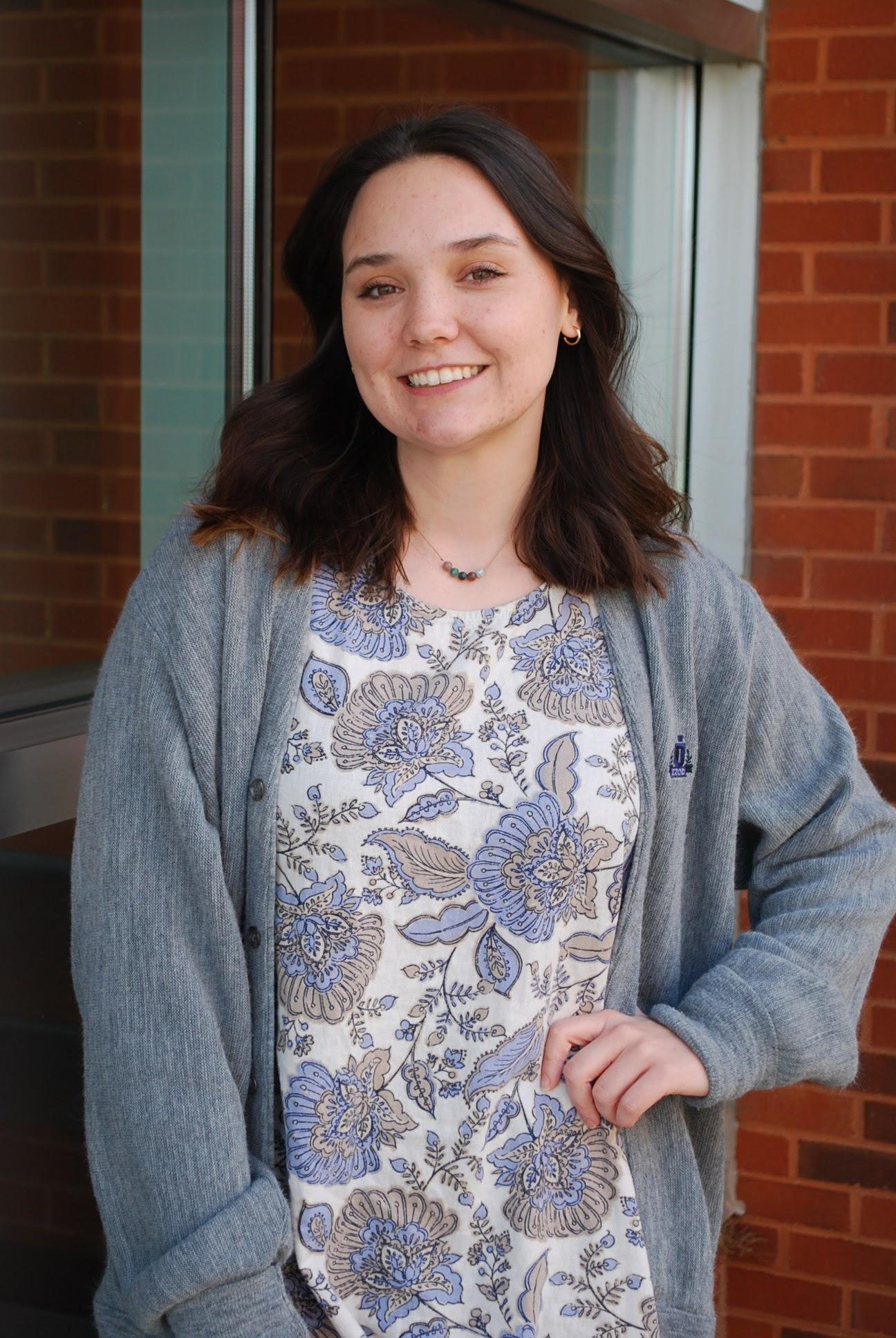 Kendall Robertson, a white woman with shoulder length dark brown hair, wearing a gray and blue patterned shirt with a gray cardigan over top. She has her left hand on her hip and is standing in front of a brick building and window.