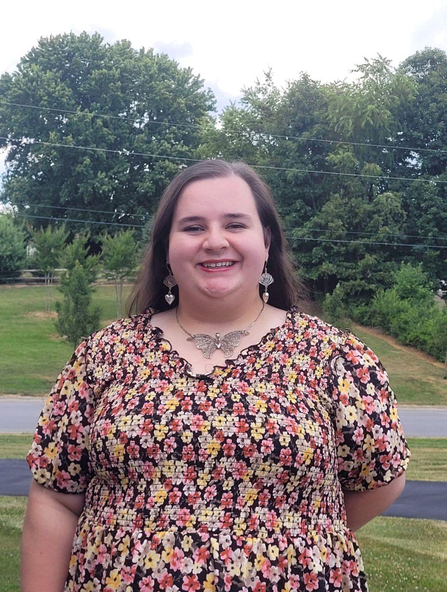 Landon Tipton, a white woman with long brown hair, wearing a red and yellow floral pattern dress with a large butterfly necklace and heart earrings. She is standing outside in front of a small tree-covered hill in the background. 