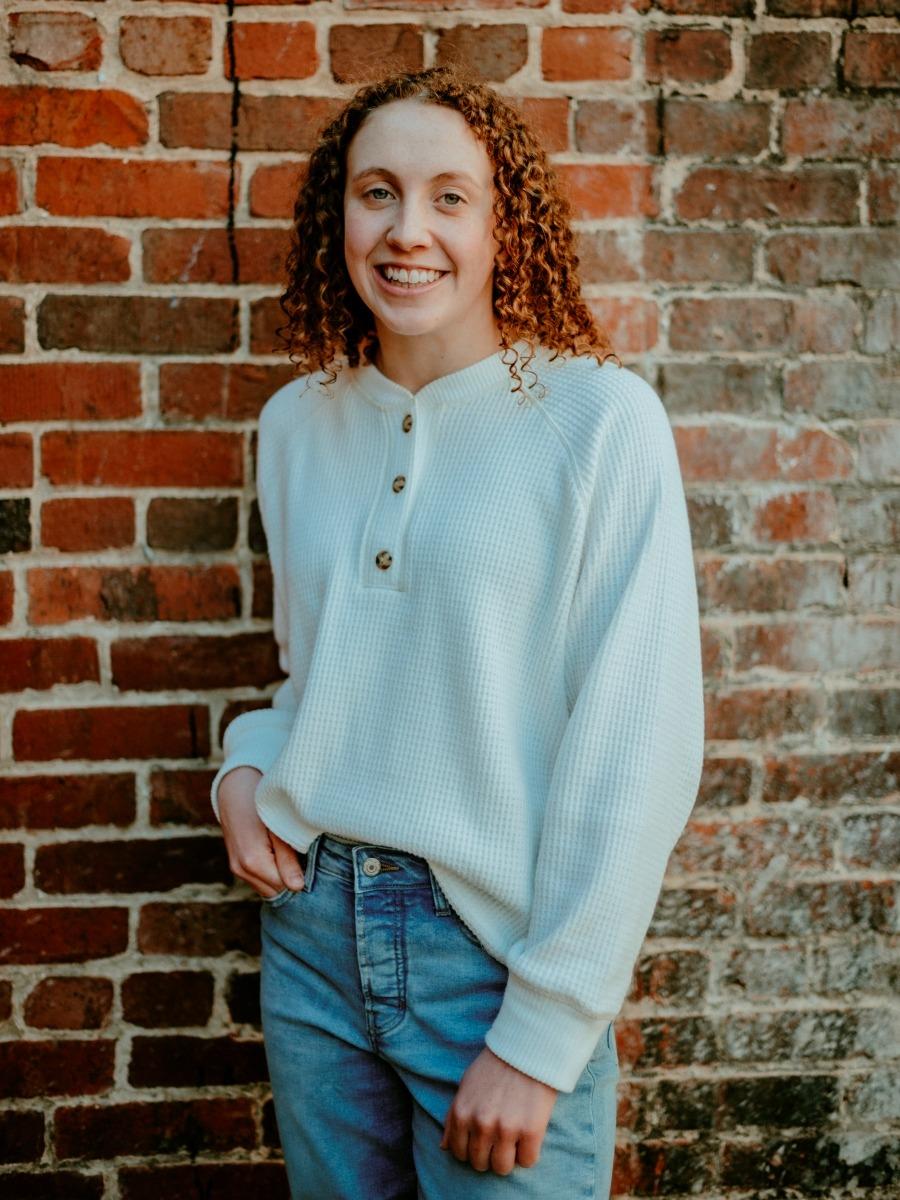 Anna Johnson, a white woman with tight curly red hair, wearing a light blue henley shirt and jeans. She is standing in front of a weathered brick wall with her right hand lightly in her pants pocket. 