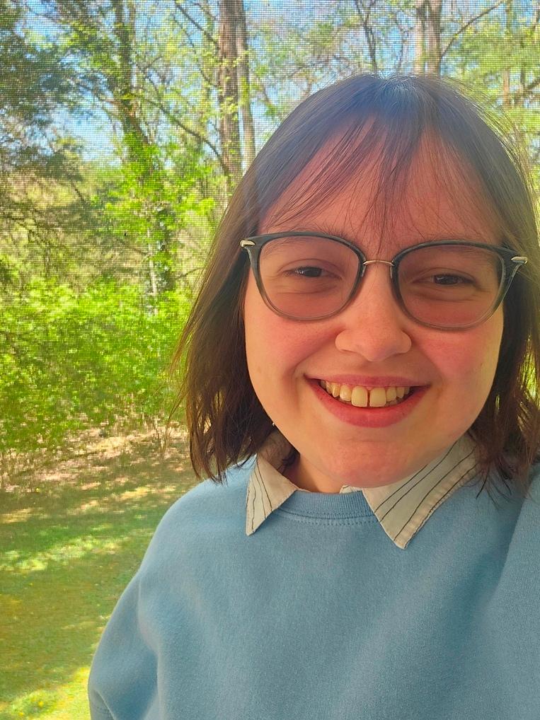 Bethanie Haga, a white woman with brown hair in a chin length bob style, wearing a light blue overshirt with a striped collar shirt underneath. She is standing under trees in the shade. 