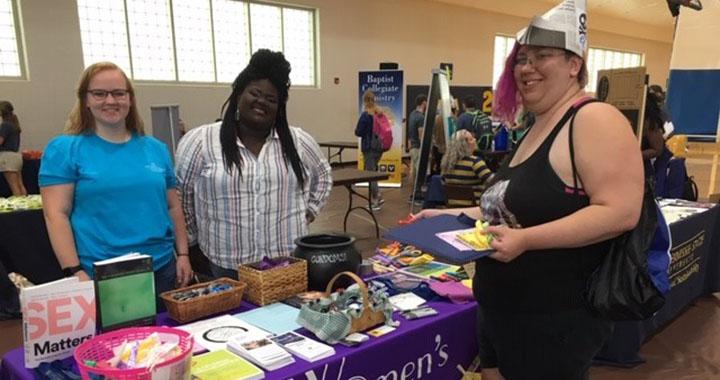 two women stand behind a table talking with another woman in front