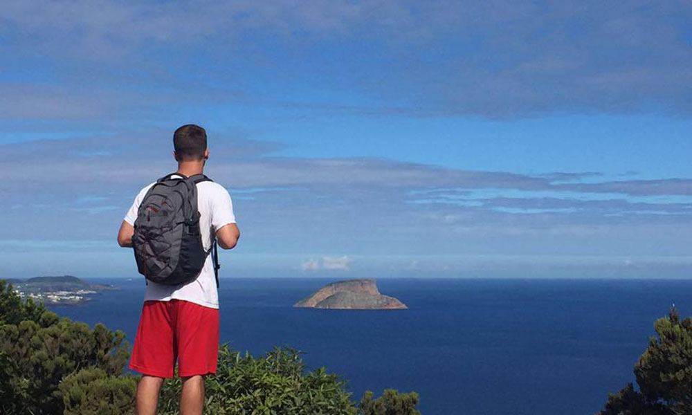 A student (Josh Bacon) stands with his back to the camera, staring out over the open ocean and island landscape.