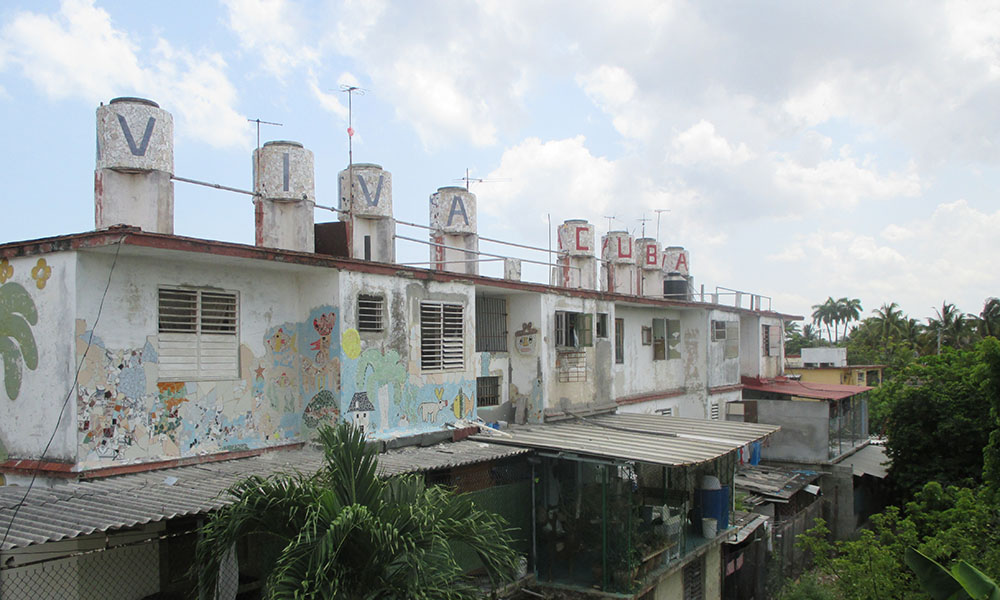 A rustic building covered in chipping white paint and homemade frescos/mosaics surrounded by tropical trees. The 8 smoke stacks have a single letter painted on them, spelling out, "VIVA CUBA."