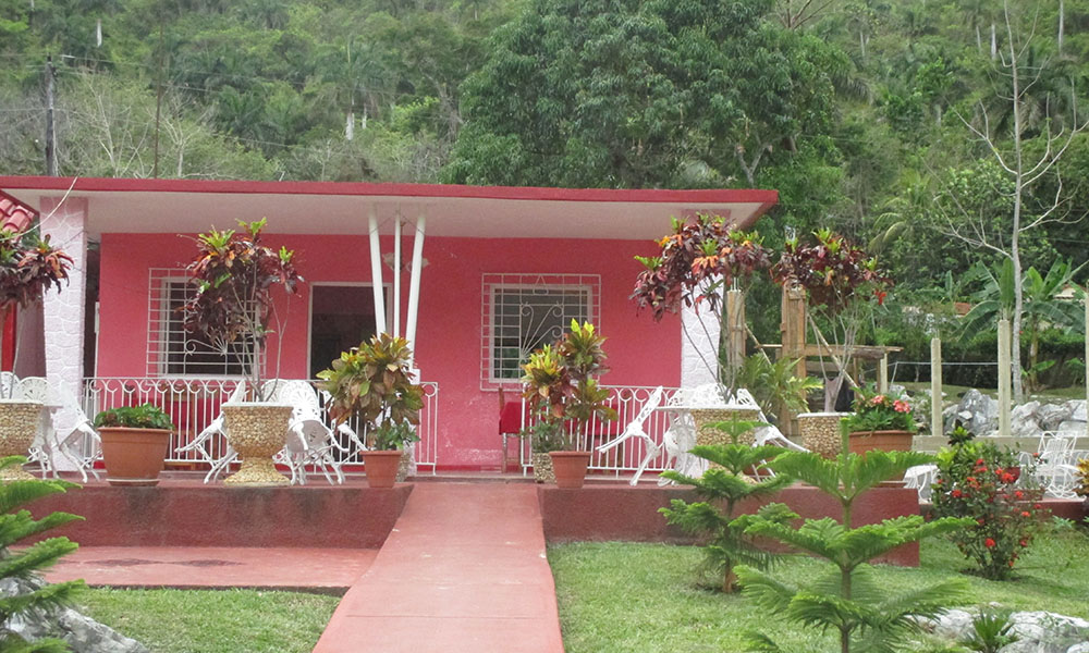 A bright pink bungalow surrounded by a gated yard, tropical trees beyond the gate in the background.