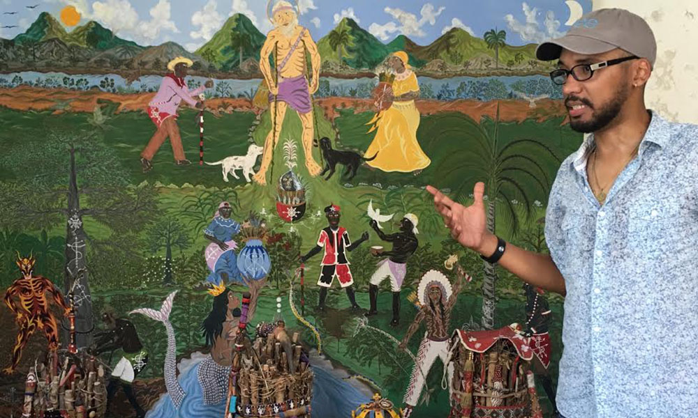 A Black man stands in front of a local street fresco of Afro/Indigenous Cuban religious/historic depictions.
