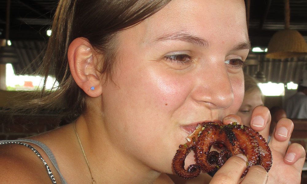 A white, female college student takes a bite out of a roasted octopus, tentacles dangling out of her mouth and over her fingers.