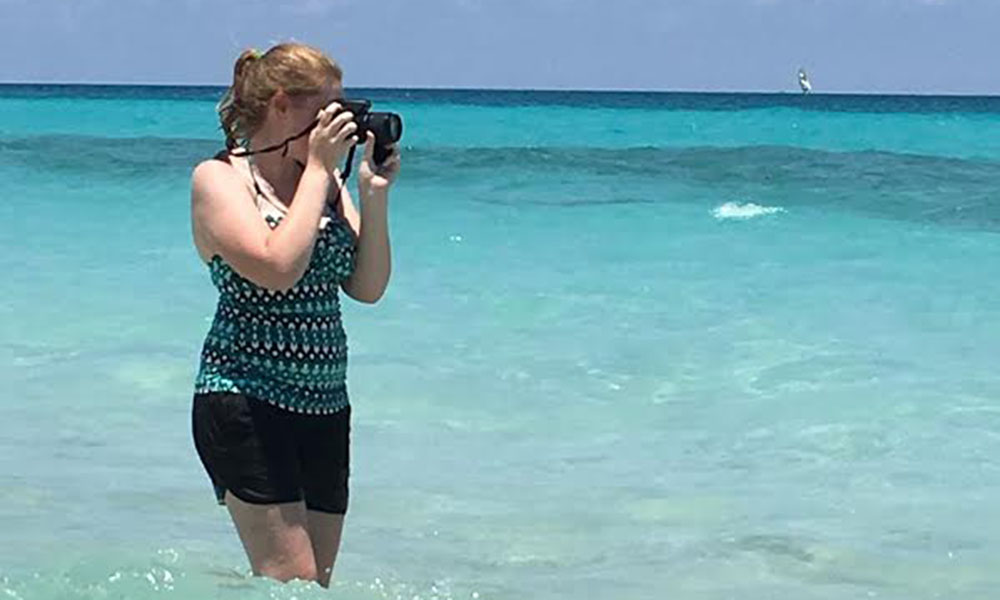 A red-headed white woman stands in the crystal clear surf, taking a photograph across frame.