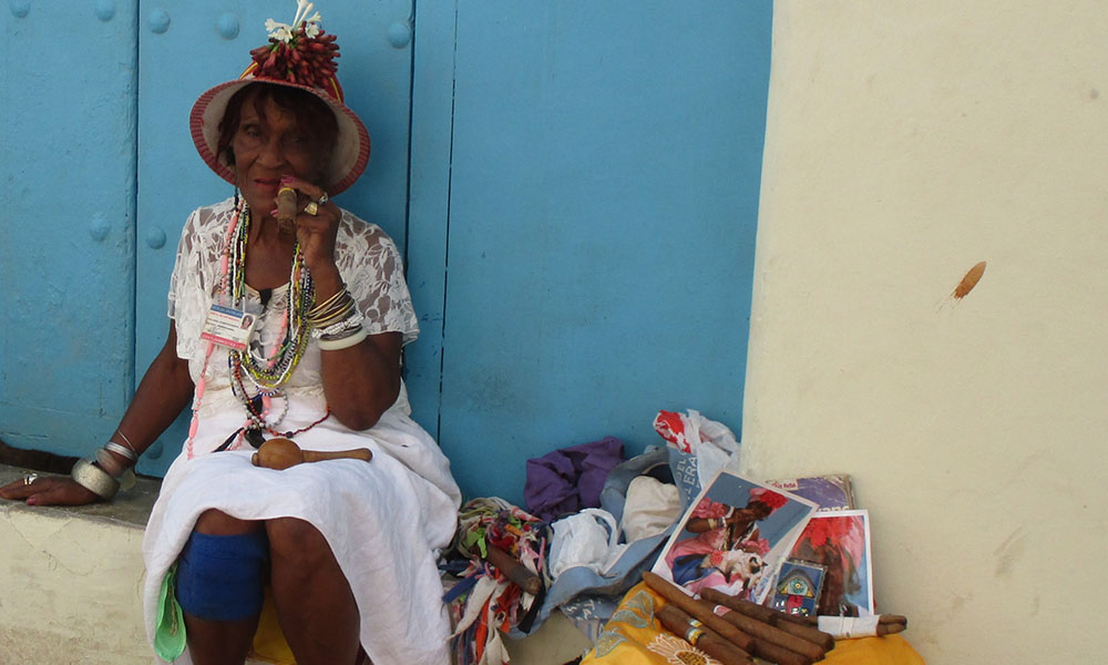 An elderly Afro-Cubana woman in a hat sits smoking a cigar, with a  basket full of photos and cigars next to her.