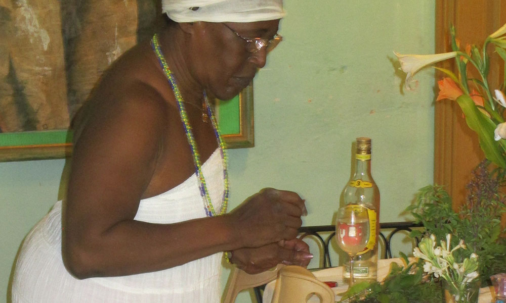 An elderly Afro-Cubana woman stands in front of a table with flowers and a glass/bottle of rum. Her crutches lean against the table in front of her.