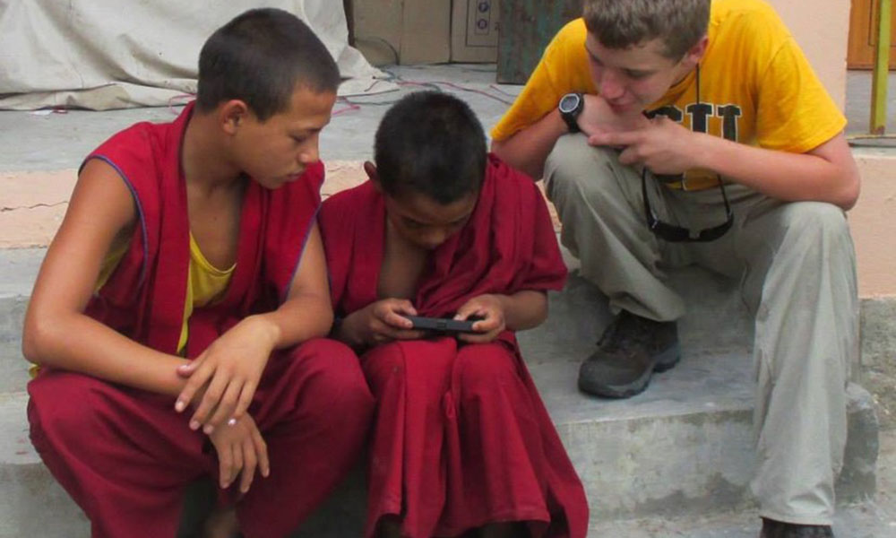 Two young local boys, monks in traditional robes and a young white, male student are seated on steps, leaning over a smartphone.
