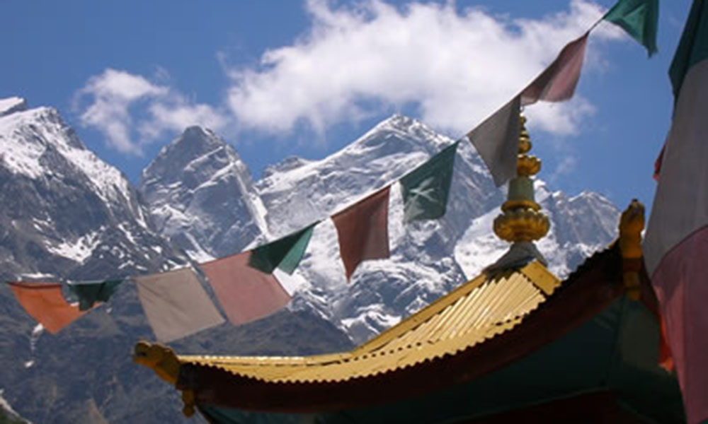Prayer flags stretch across the fram, in front of a temple roof. Snowy, Himalayan mountain peaks in the background.