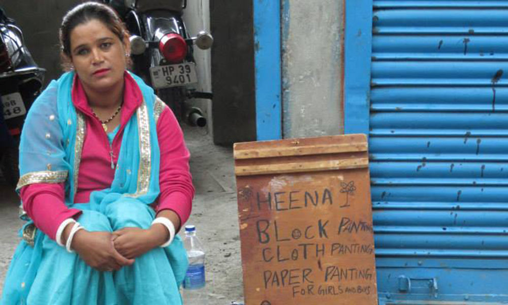 A South Asian woman sits next to her sign, advertising block Henna tattoos.