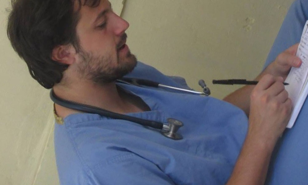 A young, white male sits in scrubs, a stethoscope around his neck, taking notes.
