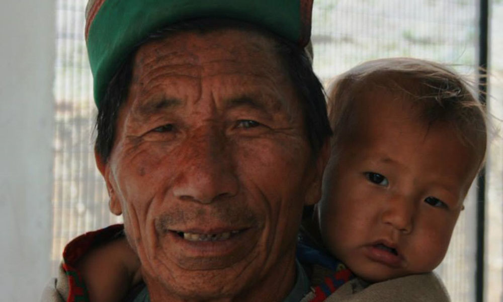 An older local man in traditional garb looks at the camera, an infant on his back.