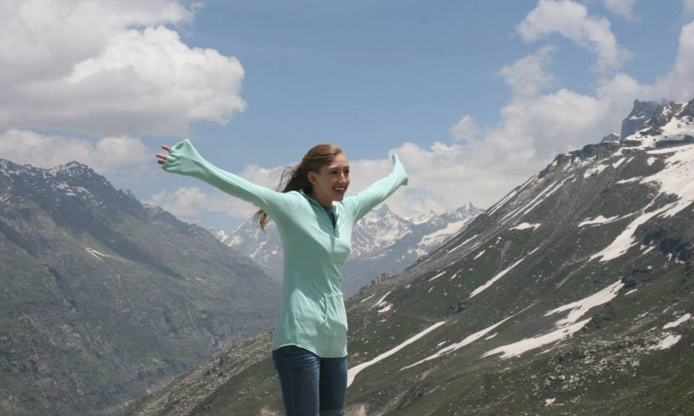 A young white woman stands with her arms stretched wide, framed by two mountains.