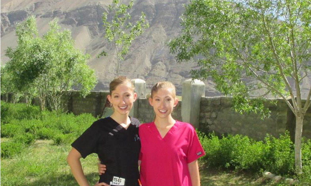 Two white women, twins, in scrubs pose in a yard fenced by a stone wall, a mountain in the background.