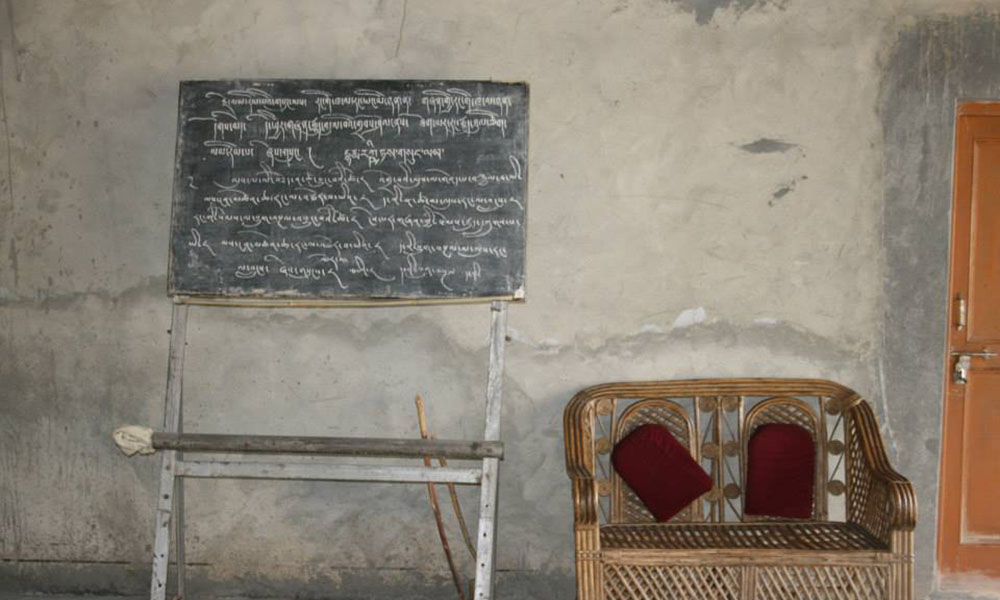 A wicker wide-seated armchair sits next to a blackboard covered in (Sanskrit?) writing.