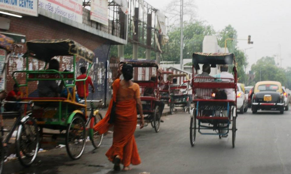 A South Asian woman in a vibrant, orange sari walks down a street, busy with rickshaws and cars.