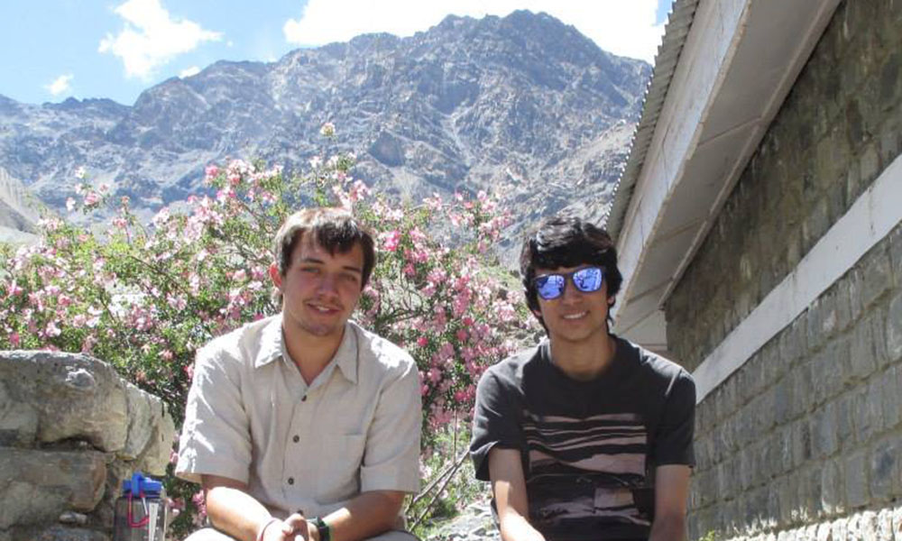 Two young men sit side by side on a stone wall, mountain and blooming trees behind them.