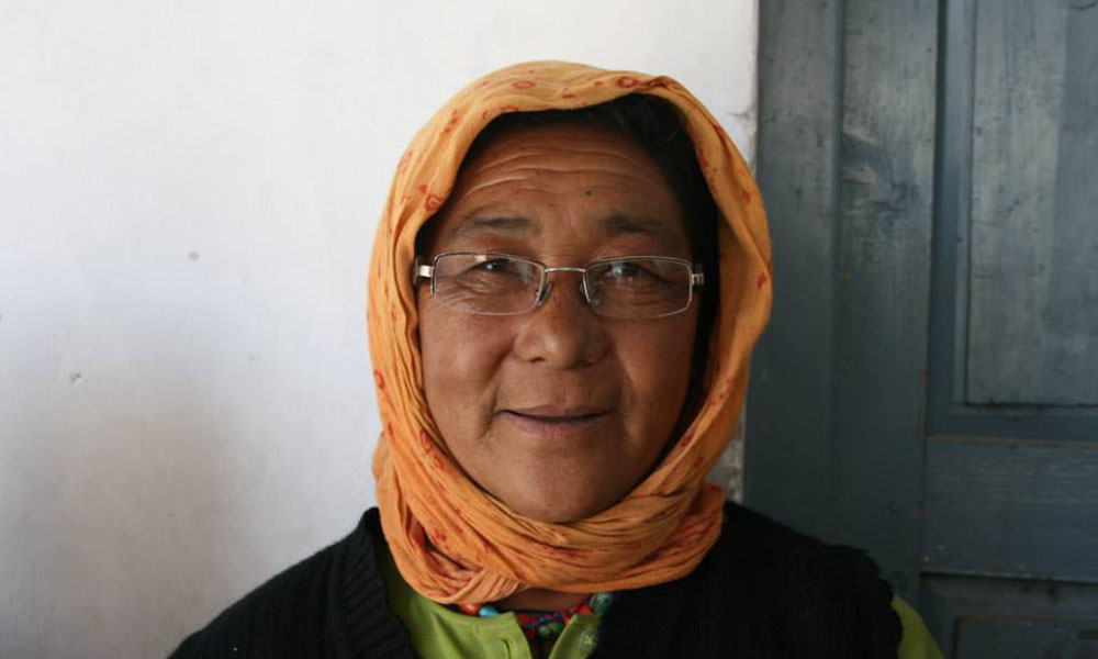 An elderly Himalayan woman in a head scarf and glasses stares directly into the camera.