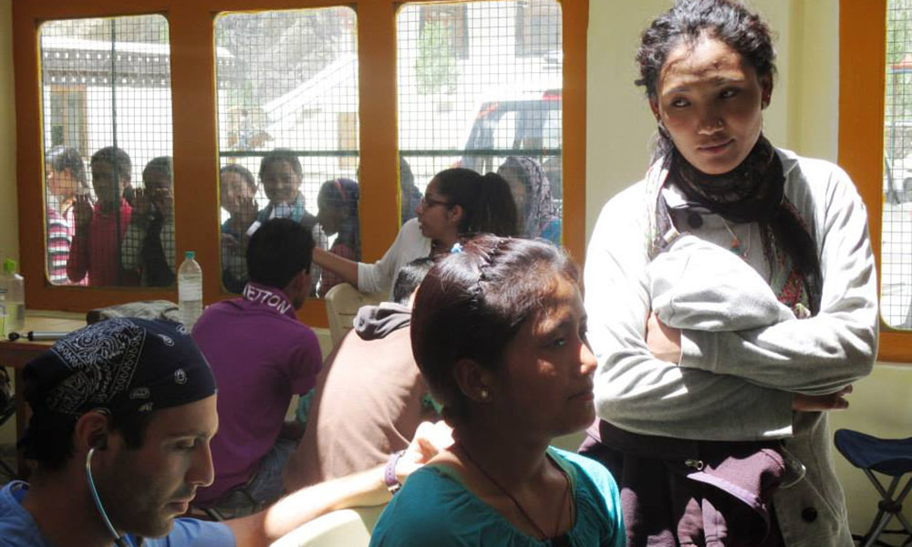 A young, male doctor crouches, checking the lungs of a Himalayan woman as her friend/relation looks on next to her. Several local children peer in through three large windows in the background.
