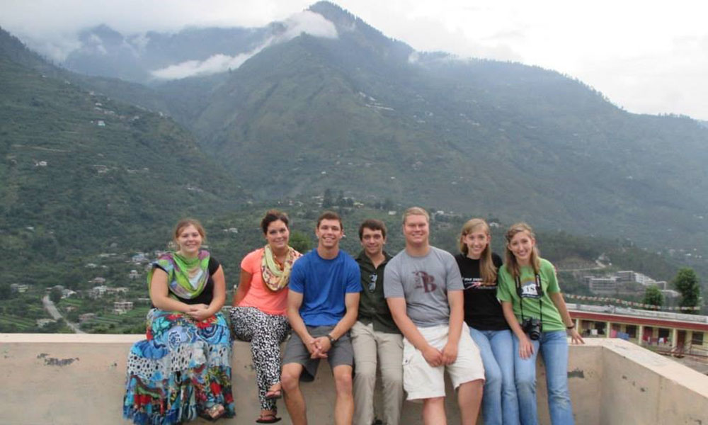 A group of students sit on a rooftop ledge, the mountains and town visible in the distance.