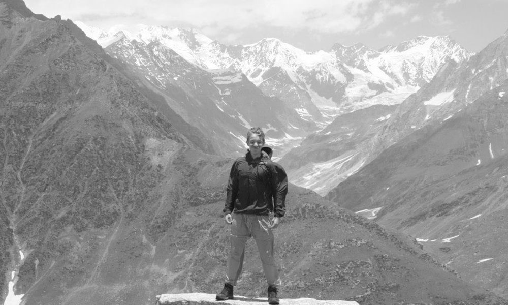 B&W photo of a young, white male college student standing in the wind, snow capped mountains looming behind him.