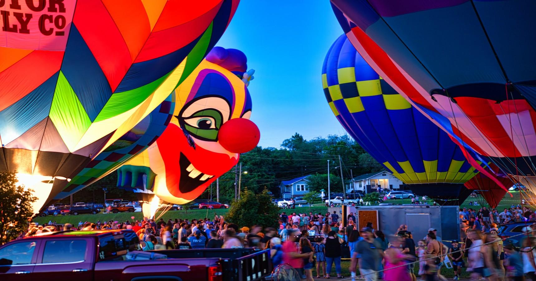 A group of hot air balloons are in a park for an after dark illumination. When the gas is turned on to inflate the balloons, they glow from within and show their colorful designs. One of the balloons is shaped like a clown’s head.