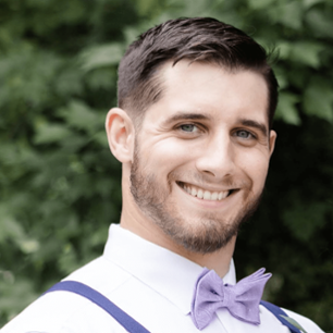 brunette man in purple bow-tie and suspenders smiling at camera