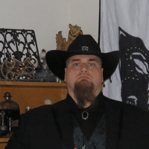 A man with a long van dyke style beard wears a black cowboy hat, staring direclty into the camera.