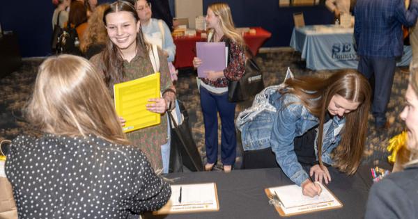Female students standing behind table recieving folders from another woman on other side of table.  