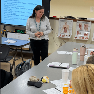 Female teacher standing in front of classroom board titles "Nutrition Label Activity." Elementary students are sitting at tables sorting Cheetos into rows. 