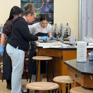 Two female highschool students, along with their teacher, lean over a science lab table looking at a fetal pig. 