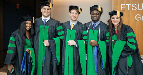 Five medical students wearing black and green doctoral regalia and tam-style caps stand side by side indoors, smiling at the camera during a hooding or commencement ceremony.