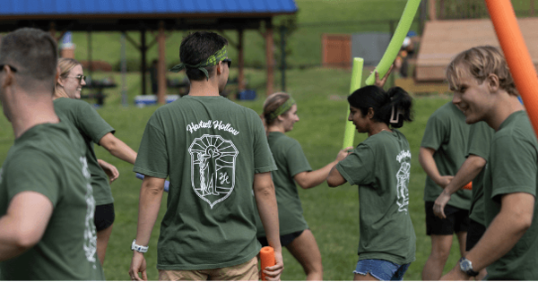 Students wearing matching green T-shirts participate in an outdoor team-building activity on a grassy field. Several students hold colorful foam noodles while interacting in small groups, with a pavilion and open green space in the background.