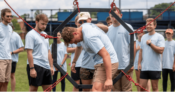 Students wearing matching light blue T-shirts participate in a team-building challenge on a grassy field. One student carefully steps through a suspended hexagonal frame while classmates stand nearby watching, encouraging and waiting for their turn.