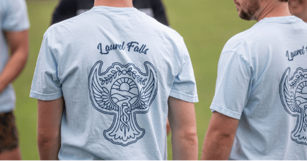 Two students stand outdoors wearing matching light blue T-shirts with a "Laurel Falls" design printed on the back. Other students are visible in the background during an outdoor team-building event.